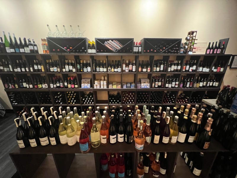 A large black shelf along a wall is filled with rows of wine bottles. A table in front of the shelf is also filled with rows of wine bottles. Photo by Alyssa Buckley.