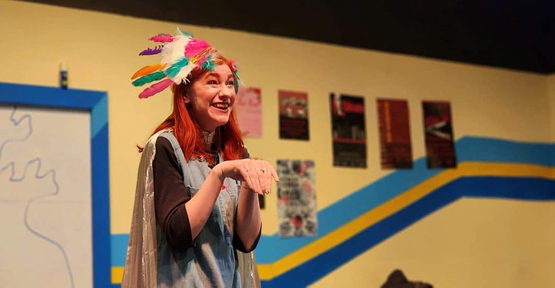 Photo of a young woman with long red hair wearing a craft-made Native American headdress in a classroom.