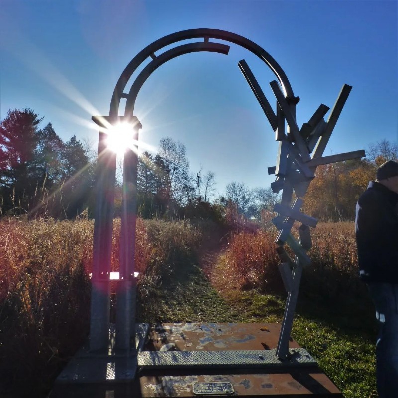 A tall metal sculpture, shaped like an arch, sits at the end of a grassy path. The sun is shining through it. Photo from Urbana Park District Facebook page.