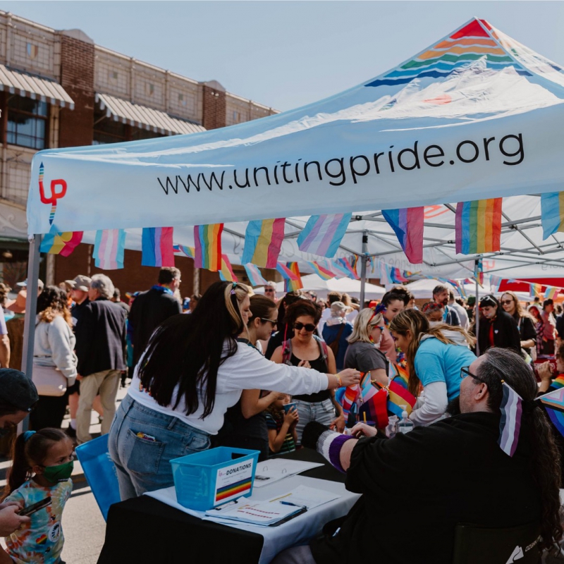 A crowd of people are gathered around a table at Pride Fest. There is a white Uniting Pride tent over the table. Photo from Uniting Pride Facebook page.