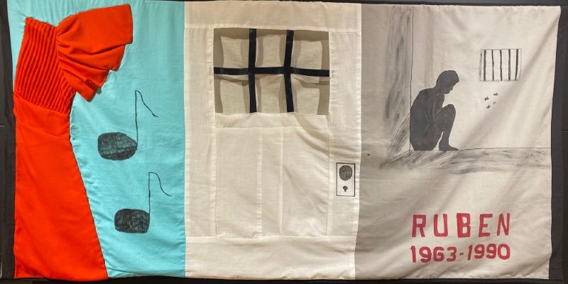 Close up of a quilt panel with three sections: one has part of a red dress and two black music notes. The center section has the door of a jail cell. The last section has the silhouette of a man in a jail cell, and says Ruben, 1963-1990 in red block letters. Photo by Julie McClure.