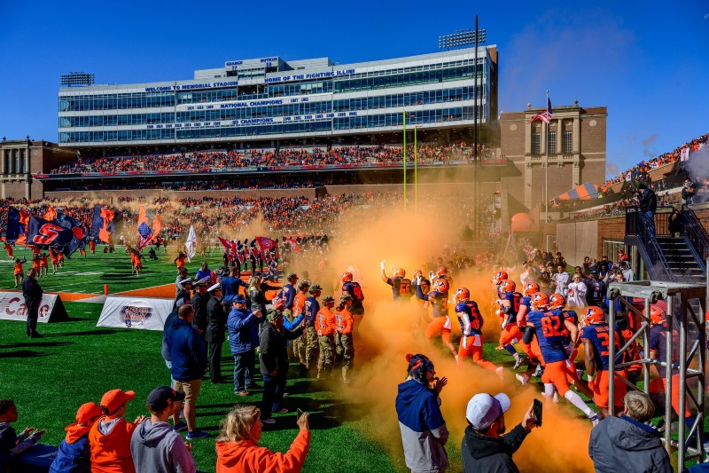 U of I football players are running onto the field through orange smoke. The cheerleaders are running ahead of them holding large blue and orange 