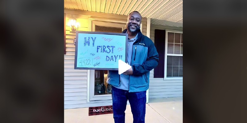 A screenshot of from TikTok of a Black man in a blue jacket standing on a front porch. He's holding a sign that says My First Day. Screenshot from TikTok. 