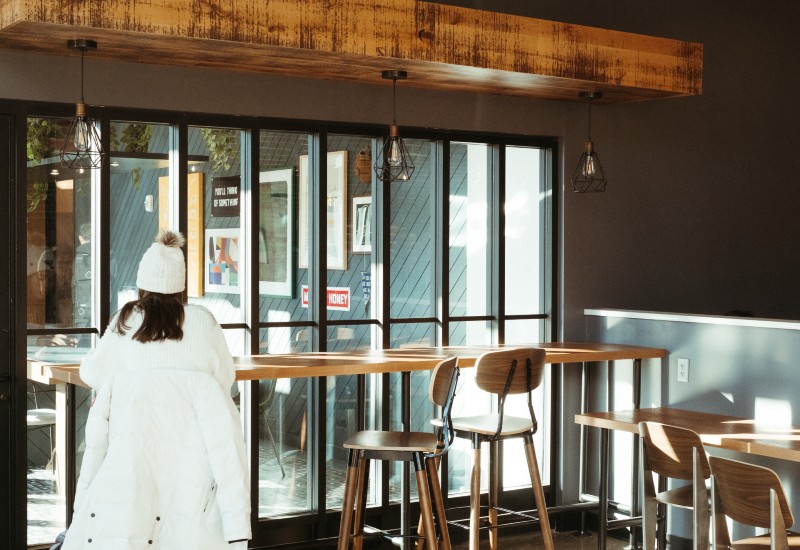 A wall of windows divided into rectangular sections. In front of the windows there is a long wooden high table with chairs. A woman in a white coat and stocking cap is sitting on one end. Photo by Patrick Singer.
