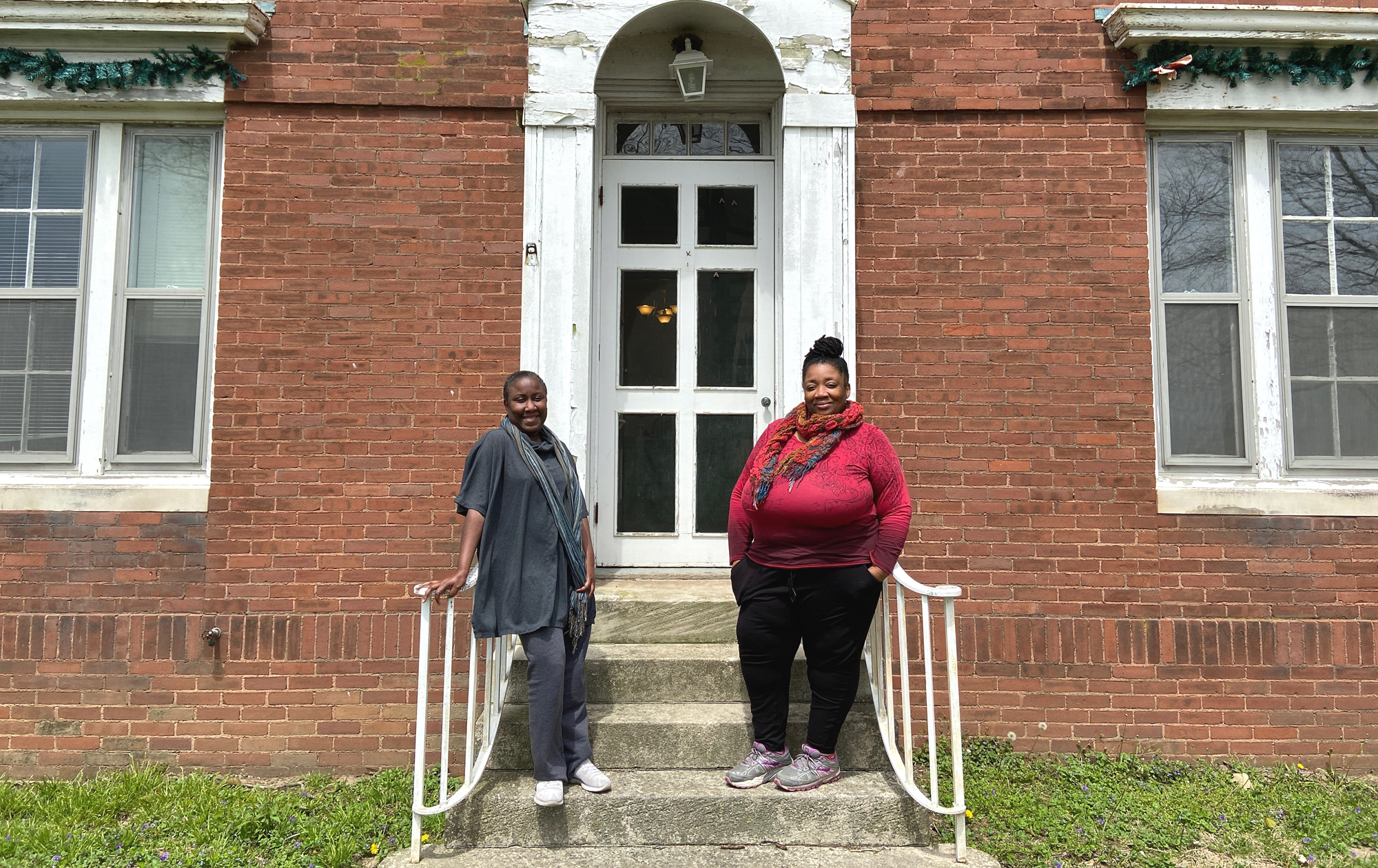 Latrelle Bright and Nicole Anderson-Cobb stand on the front steps of the artist-in-residency house at Allerton Park. Bright is on the left, wearing dark gray, and Nicole is on the right, wearing a red top. They are both looking toward the camera and smiling. The house is brick, the door is white with six window panes. Photo by Jessica Hammie. 
