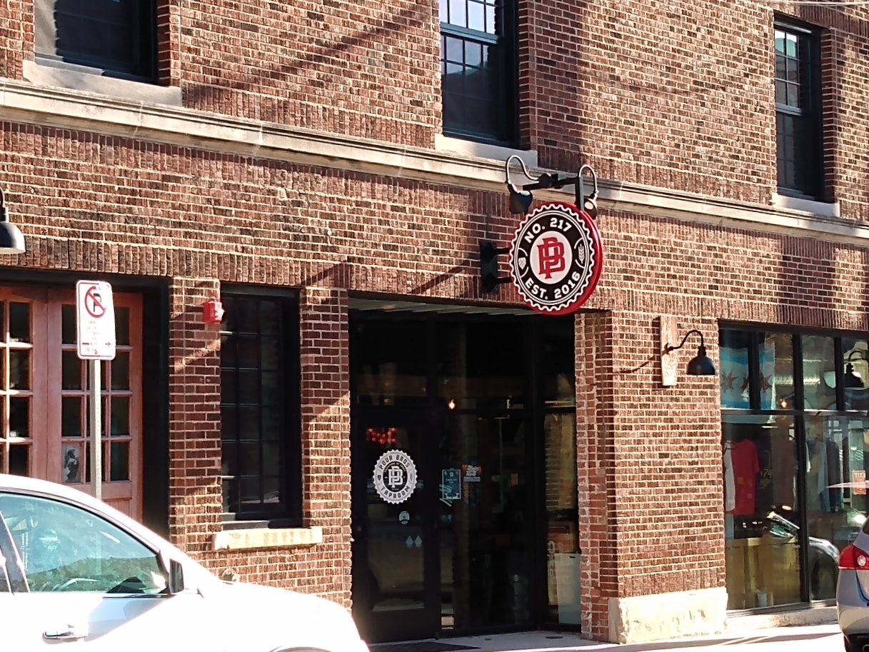 The entrance to Pour Bros. from North Market Street. A black circular sign with white lettering is mounted on a redbrick building. The same logo is also printed on a glass door. Photo by Michael O'Boyle.
