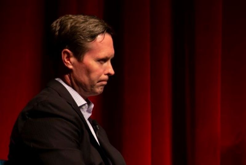  A close up of the profile of a white man with brown hair, wearing a white collared shirt black suit jacket. He is frowning. There is a red velvet curtain in the background. Photo by Steven Pratten.