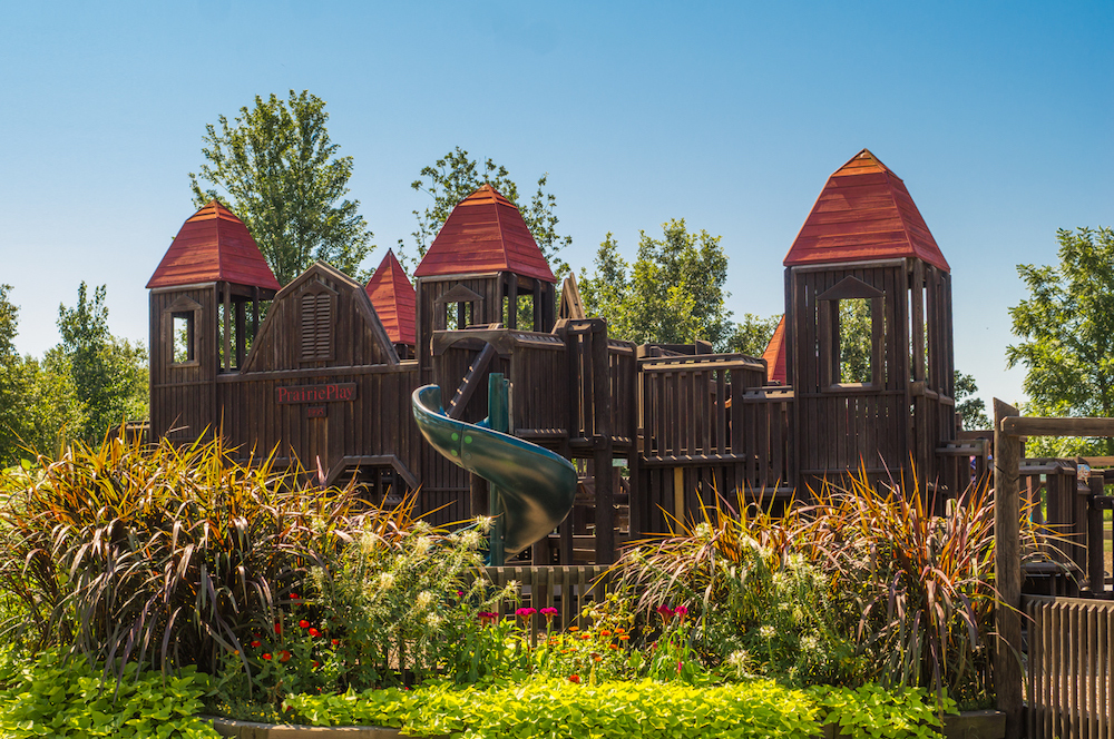 A colorful photograph of Prairie Play, the playground at meadowbrook park. Green shrubbery surrounds the playground. The wooden structure features a green twisty slide and red wooden roofs over areas of the playground. Photo from Urbana Park Districtâ€™s website.