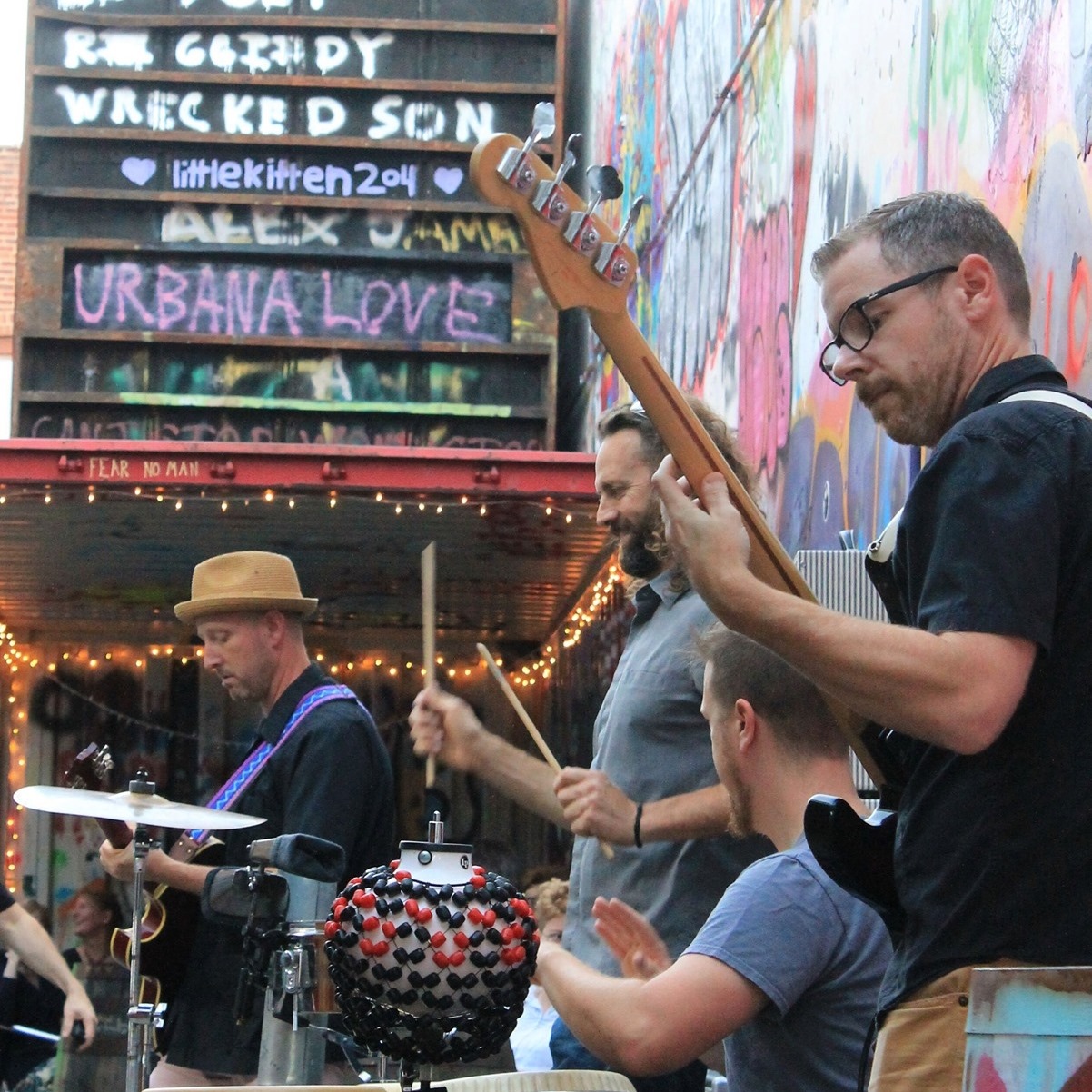 Four piece band Los Guapos perform in the Sipyard alleyway, with graffiti covered walls surrounding them. They each play their respective instrument. 