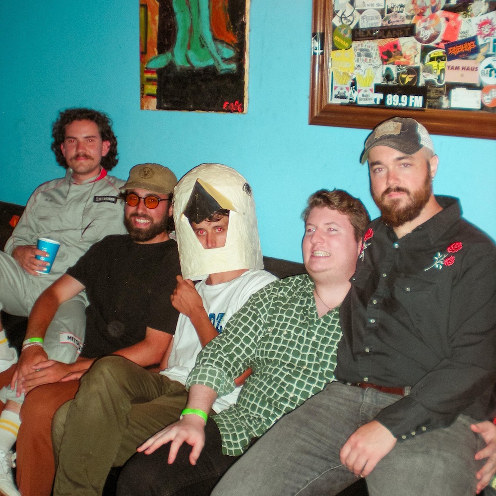 Five piece band Motel Breakfast sits on a couch smiling into the camera. The member in the middle wears a paper mache bird head.